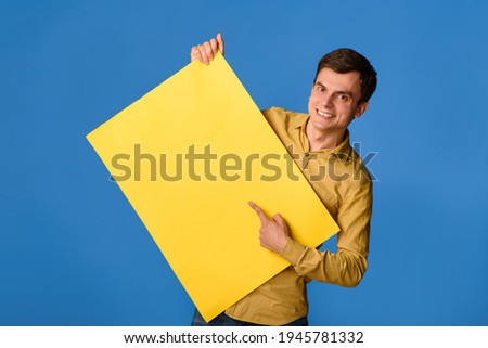 Young happy man is holding an empty yellow banner poster on blue background and laughing. Copy space for you text