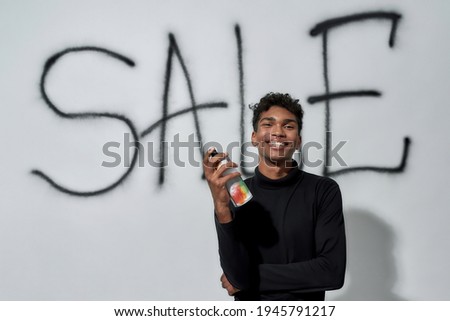 Smiling young african american man holding paint sprayer while standing near graffiti on white wall background and looking at camera. Discount and sale concept