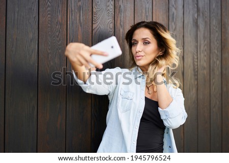 Attractive millennial hipster girl clicking selfie photos on smartphone web camera while posing in city, Caucasian female blogger taking pictures for sharing vlog content in social network