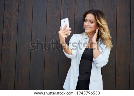Millennial woman using front mobile camera for clicking selfie pictures near copy space for advertising text, youthful female blogger shooting video vlog during leisure time at publicity area