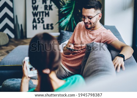 Cheerful hipster guy in optical spectacles enjoying weekend leisure with girlfriend using cellphone device for checking status of online booking for together pastime, happy couple communicate in home Royalty-Free Stock Photo #1945798387