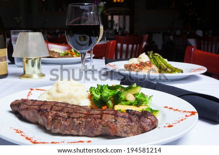 A white plate holds a delicious grilled New York Strip Steak with a side of mashed potatoes and fresh mixed vegetables.  A glass of red wine completes the meal.   Royalty-Free Stock Photo #194581214