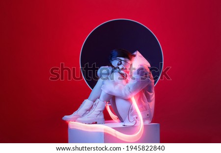 Beautiful woman in futuristic costume over red background. Girl in glasses of virtual reality. Augmented reality game, future technology, AI concept. VR. Neon light.