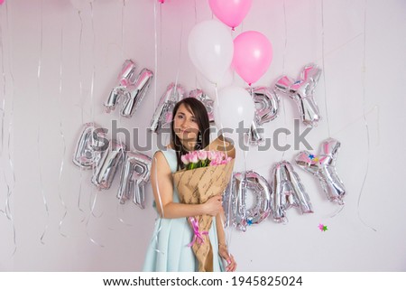 Brunette woman in mint dress holding bouquet of tulips. Birthday decorations with white and pink color balloons, bouquet of tulips and confetti for party on a white wall. Happy birthday anniversary.