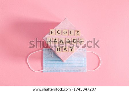  Gift and mask. Gift box and medical mask with a fools day message made from wooden cubes on a pink background, top view close-up.