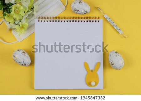 
Easter eggs with mask, blank notepad, thermometer, bunny and flowers lie on a yellow background, close-up top view.