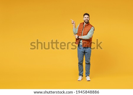 Full length young smiling happy confident smiling cheerful fun caucasian man 20s in orange vest mint sweatshirt point index finger aside on copy space area mock up isolated on yellow background studio