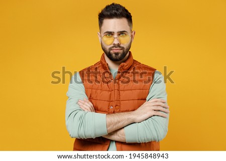 Young serious confident shrewd caucasian man 20s in orange vest mint sweatshirt glasses look camera hold hands crossed folded isolated on yellow background studio portrait. People lifestyle concept.