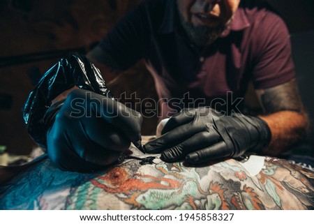 Tattoo artist wearing black gloves creating a picture on a mans back with a tattoo machine