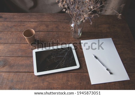 Modern office workplace with tablet pc on the table. On the table is a tablet computer with a sheet of white paper, dried flowers in a glass and a disposable cup of coffee.