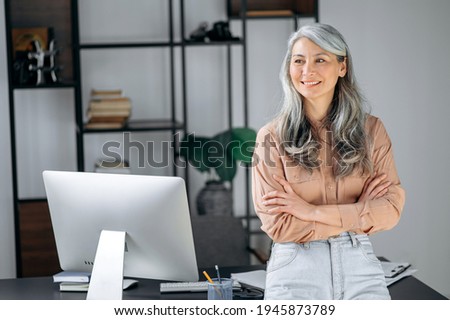 Portrait of a pretty influential successful gray-haired Asian woman, business leader, manager, ceo stands in a modern office, stylishly dressed, crossed arms, looking to the side, smiling friendly Royalty-Free Stock Photo #1945873789