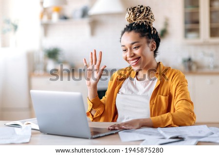 Happy successful beautiful stylish african american girl, student or freelancer, talking by video call, online meeting with colleagues or friends, greeting with hand gesture, smiling friendly Royalty-Free Stock Photo #1945875280