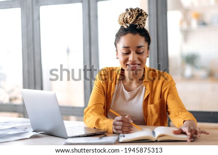 Online education. Joyful clever focused African American stylish young woman with dreadlocks, student, studying online at home, watching online video lesson, uses laptop, taking notes, smile Royalty-Free Stock Photo #1945875313