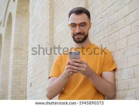 Handsome smiling man using mobile phone, communication online, chatting, watching video standing on the street. Young bearded hipster wearing stylish eyeglasses holding smartphone shopping online