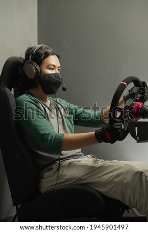 handsome Asian young man gamer wearing mask in headphones with pc computer playing car racing video game at home and steering wheel