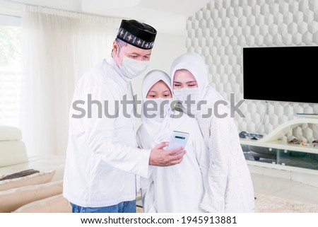 Happy parents with their daughter wearing face mask while making a video call by using a cellphone during Eid Mubarak in the living room. Shot at home