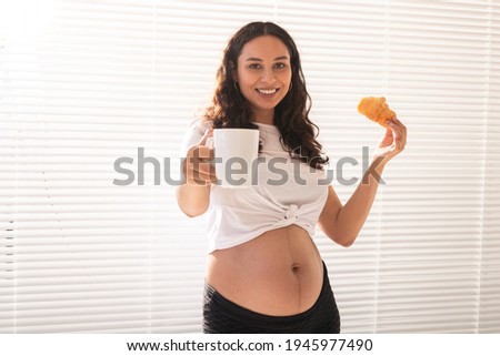 Beautiful pregnant woman holding croissant and cup of coffee in her hands during morning breakfast. Concept of good health and positive attitude while expecting baby