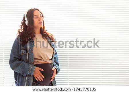 Beautiful young peaceful pregnant woman listens to pleasant classical music using smartphone and headphones. Concept of positive attitude before childbirth. Copy space