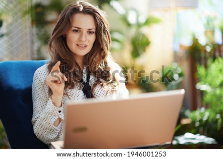Green Home. trendy woman with long wavy hair in the modern house in sunny day having web cam meeting on a laptop.