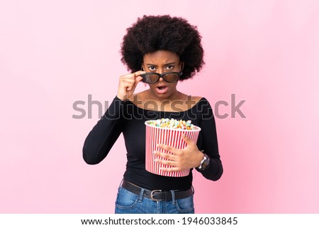 Young African American woman isolated on pink background surprised with 3d glasses and holding a big bucket of popcorns
