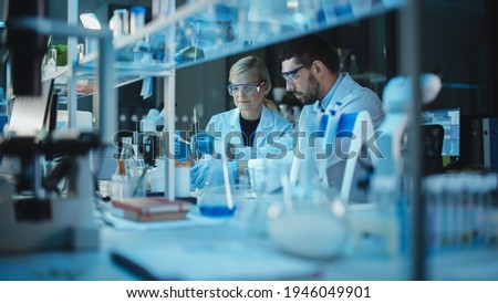 Female and Male Medical Research Scientists Have a Conversation While Conducting Experiments in Test Tubes with Liquid Samples with and in Beakers with Solid Speciments. Modern Science Laboratory. Royalty-Free Stock Photo #1946049901
