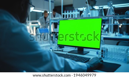 Medical Research Scientist Working on Desktop Computer with Green Screen Mock Up Template in Applied Science Research Laboratory. Lab Engineers in White Coats Conduct Experiments in the Background.