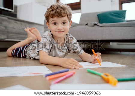 Portrait of school-age boy lies on the living room floor and paints pictures on white sheets of paper with colored markers. The boy is spending his afternoon free time at home.