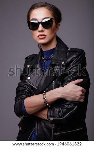 High fashion photo of a beautiful elegant young woman in a pretty leather jacket, blue dress, accessories, sunglasses,  posing over gray background. Studio Shot. Portrait