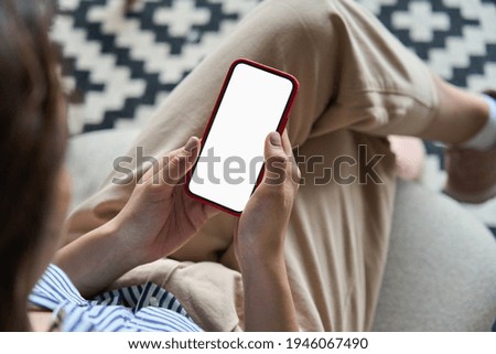 Young woman or teen girl hands holding mobile smart phone with mockup white blank display, empty screen at home. Shopping, delivery apps, social media applications ads, over shoulder close up view. Royalty-Free Stock Photo #1946067490
