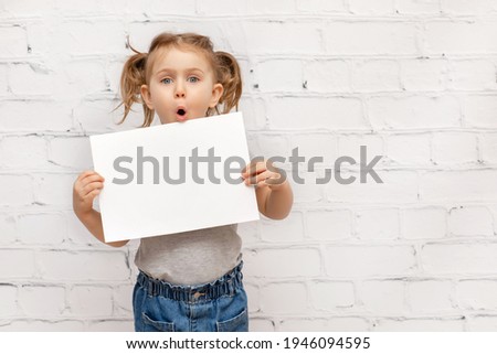 Surprised child over white brick wall holding white paper sheet with expression face.Concept of advertising goods, seasonal sale, discount. Shopping and retail. Promoting goods. Place for text. Mockup