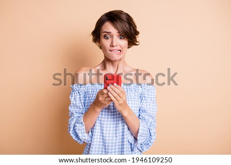 Photo portrait of scared woman biting lower lip holding phone in two hands isolated on pastel beige colored background