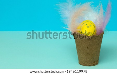 happy Easter. A yellow egg with a painted chicken in a glass with feathers on a blue background. Horizontally. Copy space.