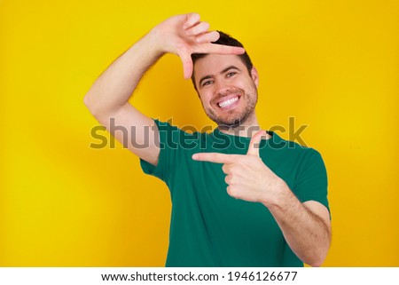  young handsome caucasian man wearing green t-shirt against yellow background making finger frame with hands. Creativity and photography concept.
