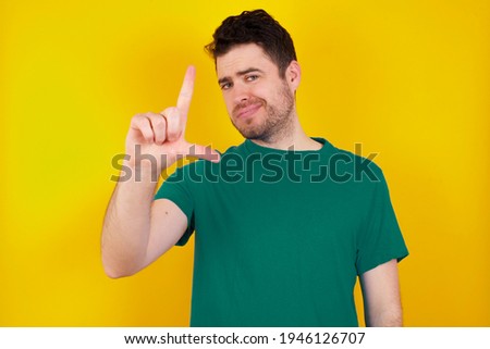  young handsome caucasian man wearing green t-shirt against yellow background making fun of people with fingers on forehead doing loser gesture mocking and insulting.