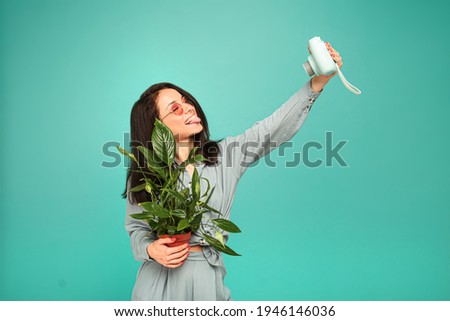 Photo of attractive lady in sunglasses holds camera, plant and take a selfie. Wears grey shirt, isolated turquoise color background.
