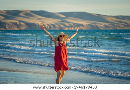 Happy attractive Mature woman in red dress enjoying outdoors and freedom on the beach, open arms outstretched with blue ocean and beautiful landscape. Retirement lifestyle and Wellbeing concept.
