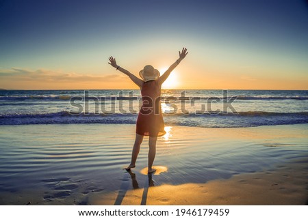 Happy attractive Mature woman in red dress enjoying outdoors and freedom on the beach, open arms outstretched with blue ocean and beautiful landscape. Travel, People, Hope and Wellbeing concept.