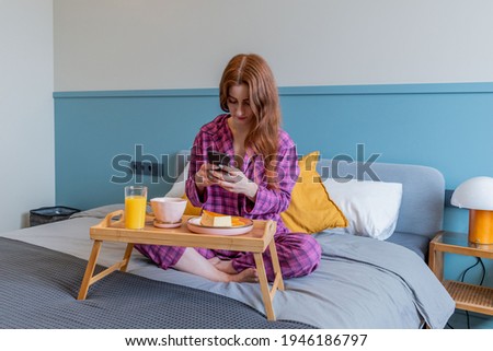 Young woman taking pictures of her breakfast while sitting in bed. Technology concept in modern world. Red-haired girl in pink pajamas