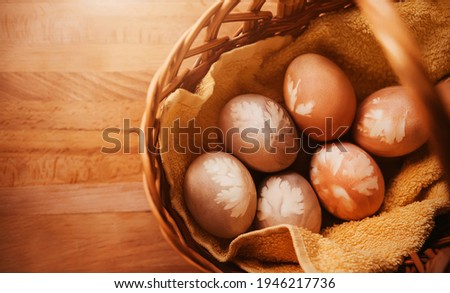 On a wooden table is a wicker basket, in which on a yellow cloth are chicken eggs, painted with natural dyes with drawings of plants. Easter decor.