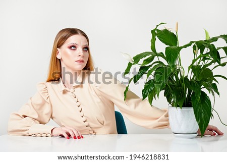 young girl in the office sits on chair. there is green flower on the table. woman has creative makeup on her face