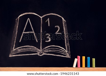 A book drawn with white chalk on a black board with a letter and numbers and colored crayons near
