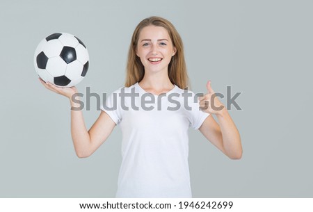 Young woman holding a soccer ball and showing thumb up, looking at the camera, isolated on gray background.