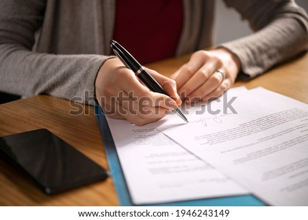 Close up of hands of young woman signing legal documents on desk. Close up of woman hand holding pen and signing legal paper seated at desk. Detail of lawyer filling official document, deal done.