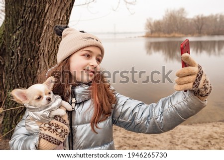 A teenage girl takes pictures of herself and the dog on a smartphone or speaks via video communication. Girl in a winter jacket and chihuahua. Girl on a background of a river with a dog. A pet.