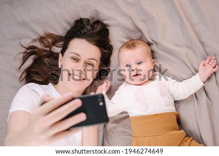 Top view of beautiful mum and baby daughter lying on bed have fun use cell phone watch cartoons, mom with infant girl making online call vlog. Parental control, modern tech, younger generation concept