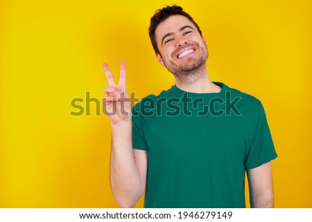  young handsome caucasian man wearing green t-shirt against yellow background smiling with happy face winking at the camera doing victory sign. Number two.