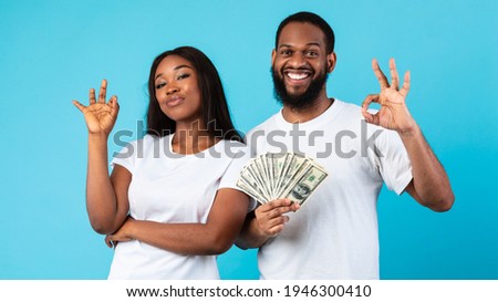 Win Concept. Portrait of confident young African American couple holding bunch of money, celebrating success together, showing okay sign gesture, posing over blue studio background, banner
