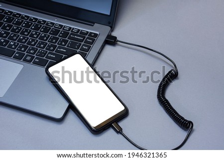 Mock-up of a smartphone with a white screen close-up being charged from a laptop on the background of a gray table