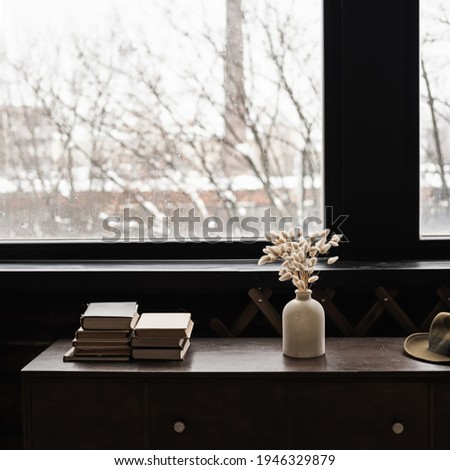 Rabbit bunny tail grass bouquet, books stack on solid wooden table against window. Aesthetic dark home interior design concept. Beautiful floral foliage composition.