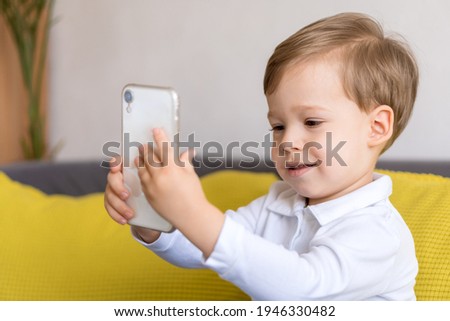 close-up happy little cute preschool baby boy talking video conference by smartphone in living room at home on yellow gray sofa with happiness smile. Childhood, online communication, tehnology concept